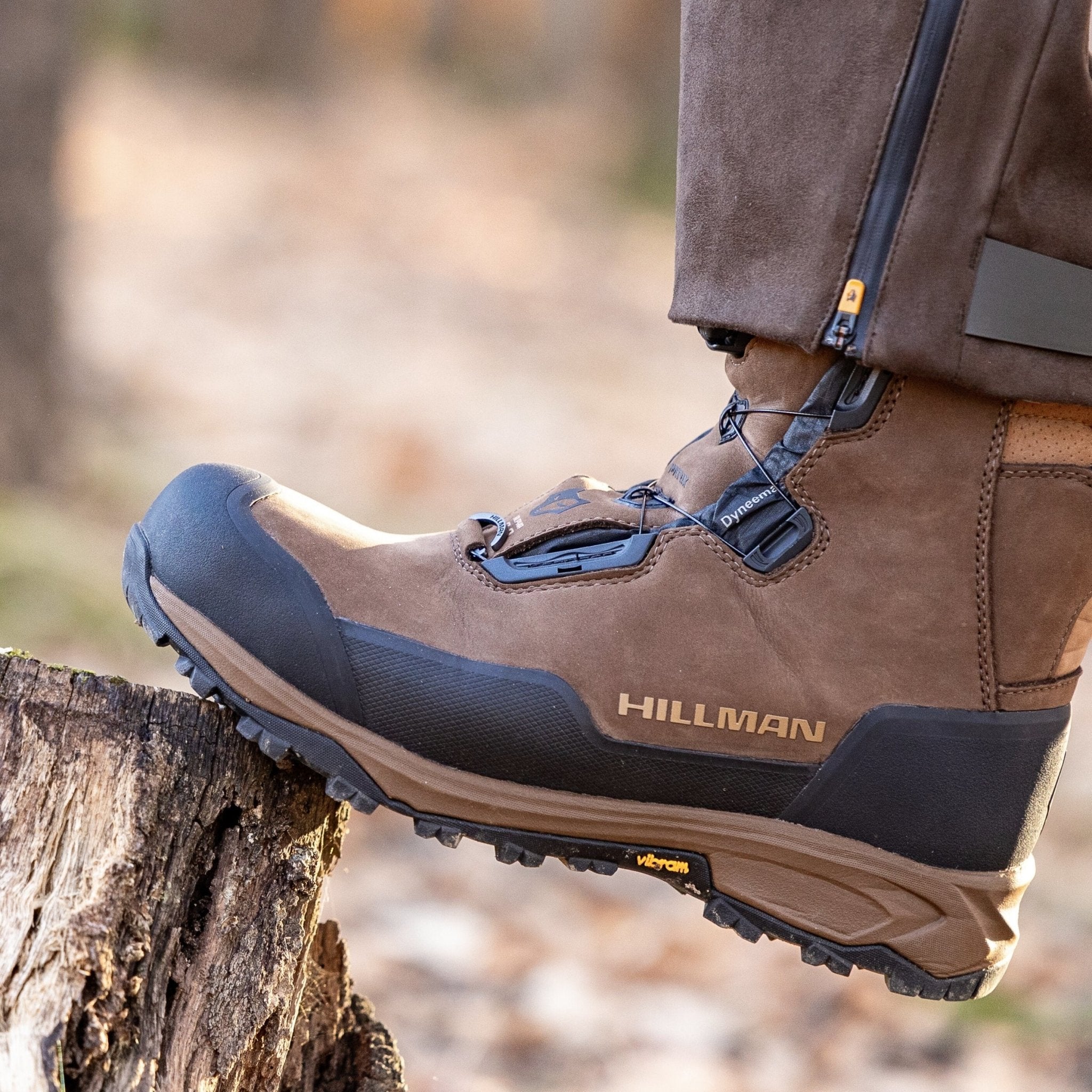 A close-up of a person wearing HILLMAN Insulated Hunting boots Aerogel™3-Season with black soles, standing on a tree stump outdoors. The boots display the Vibram logo and are paired with zippered pants.