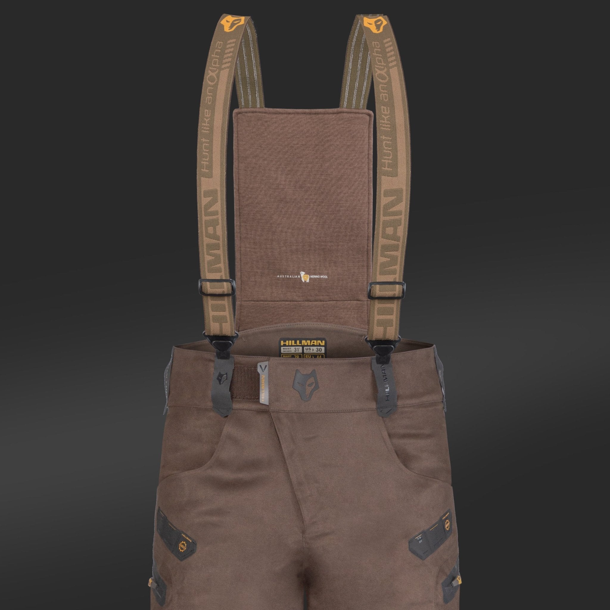 Close-up of HILLMAN GEAR Insulated Hunting Pants 6OL Cold Weather Waterproof Bibs featuring brown fabric, Matrix Insulation, dark reinforced patches, belt loops, multiple pockets, and adjustable branded suspenders. Solid black background.