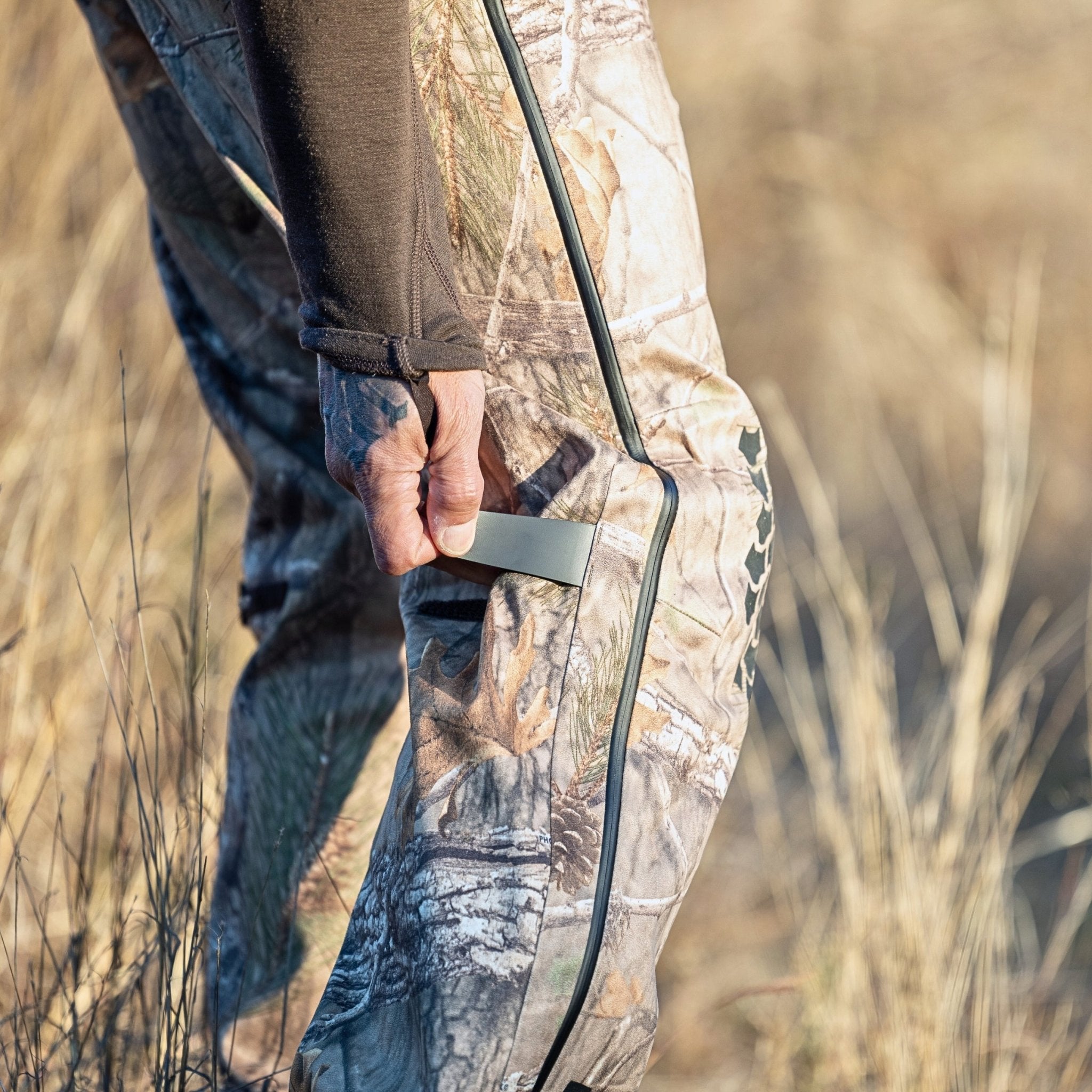 A person wearing HILLMAN GEARs 5WL Waterproof Camo Hunting Pants stands outdoors on dry, brown grass. They hold a small object in their right hand near the side seam of these ultra-lightweight camouflage trousers.